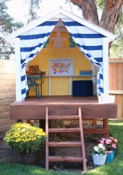 a colorful kids’ outdoor space with yellow walls and a blue and white curtain, with a deck and a staircase plus potted blooms around