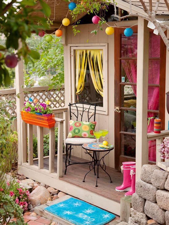 a tan planked playhouse with bold curtains, a bright garland and lamps, greenery, a bold accesories and decor