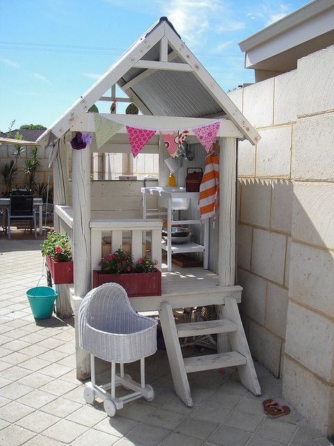 a white mini playhouse with a console table and a stool, with a ladder, a colorful garland and textiles