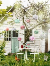a white kids’ playhouse with window frames and an aqua door, a tree with pastel pompoms and a metal dining table and chairs around