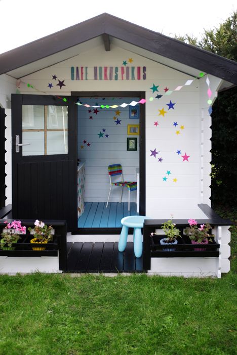 a black and white kids' playhouse with a bit of kids' furniture inside, with colorful letters and stars, with potted blooms and a bright garland
