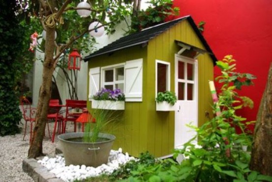 a bright green mini kids' playhouse with a black roof, white window frames and a door, with greenery and a small pond
