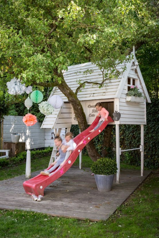 a raised white kids' playhouse with a ladder and a red slide, with potted blooms and greenery is a great spot for kids' games