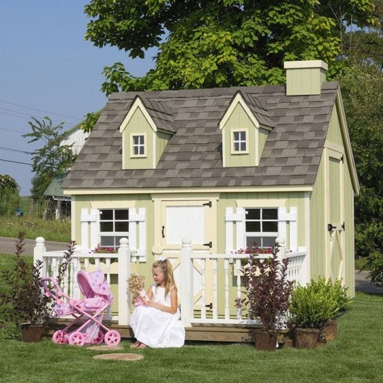 a small green kids' playhouse with a dark roof, white window frames and a door, potted plants and a pink baby carriage