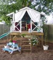 a pretty small kids’ playhouse with a staircase and a raised deck, colorful vintage furniture inside, a curtain for privacy and some potted plants