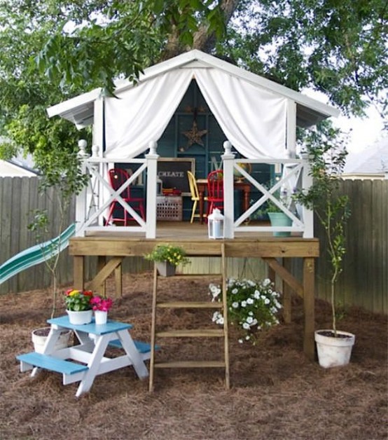 a pretty small kids' playhouse with a staircase and a raised deck, colorful vintage furniture inside, a curtain for privacy and some potted plants