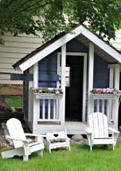 an elegant blue and white kids’ playhouse with potted bold blooms and some white garden furniture next to the house
