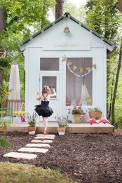 a whitewashed kid’s playhouse with a large window, some cool furniture inside, outdoor furniture and potted blooms plus a flamingo