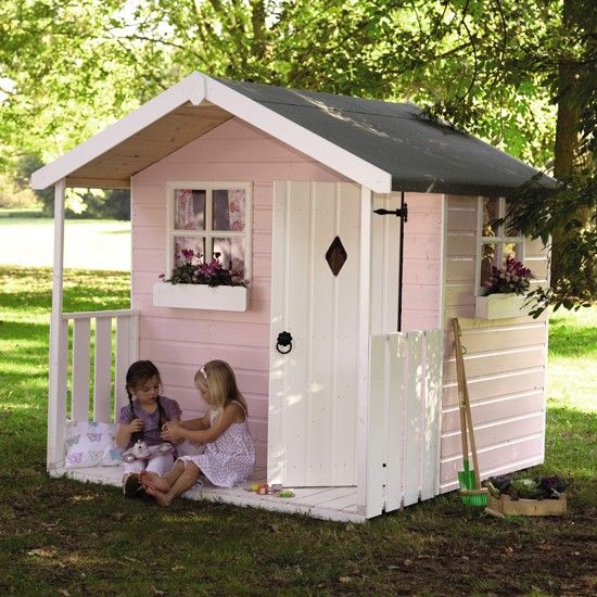 a light pink kids' playhouse with a white door and windows, with a small porch and potted blooms and gardening tools around