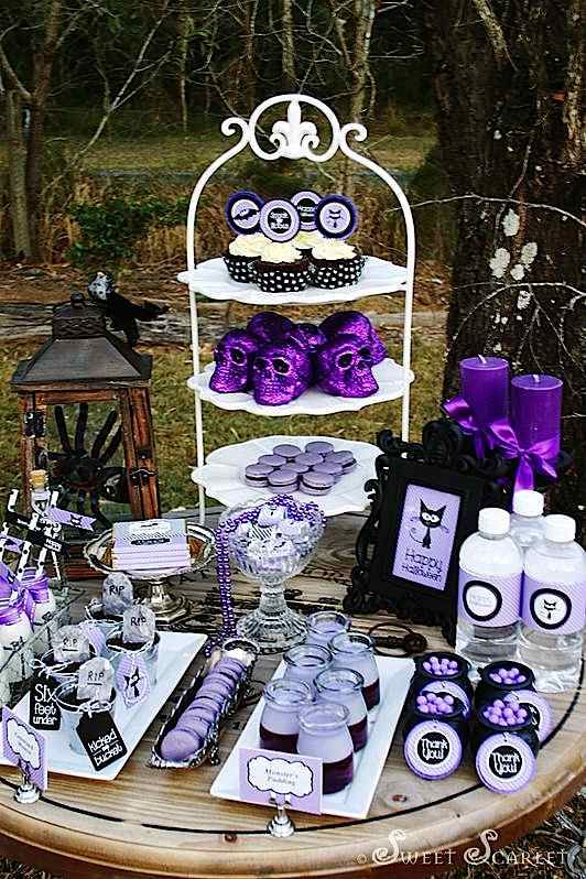 a black and purple Halloween sweets table with various desserts, candies and cupcakes, with purple glitter skull decor