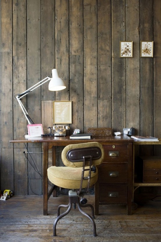 a rustic industrial home office with rough wooden walls, a vintage desk, a comfy chair and modern lamps and art