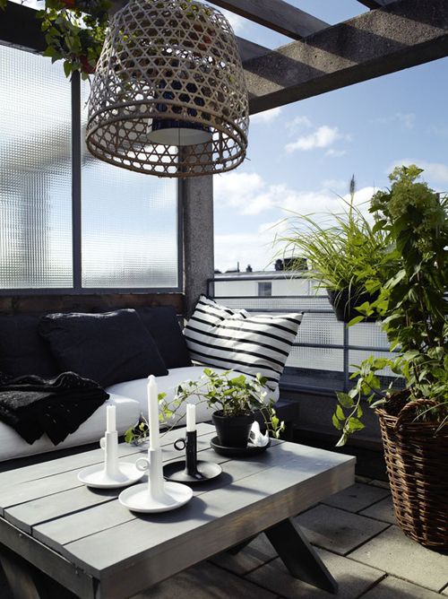 a stylish Nordic balcony with a black bench and black and white textiles, a wooden coffee table, a woven pendant lamp and some potted greenery