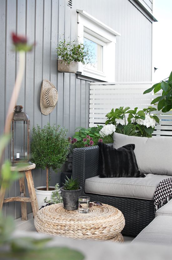 a lovely Scandinavian balcony with black wicker furniture with grey and black upholstery, a jute pouf, candle lanterns and potted blooms and plants