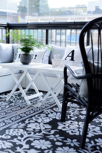 a black and white Scandinavian balcony with a printed rug, a white corner sofa, a black chair and white folding tables and greenery