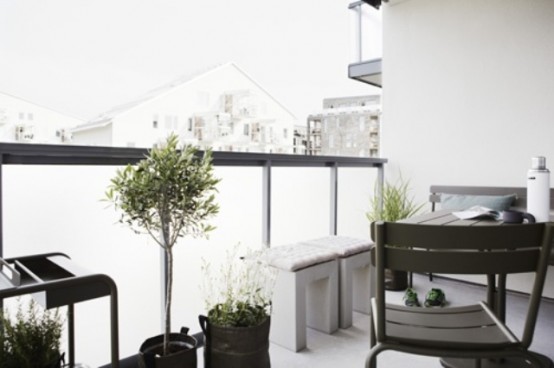 a monochromatic Nordic balcony with black and grey metal and wood furniture, white cinder stools, potted greenery and plants is welcoming