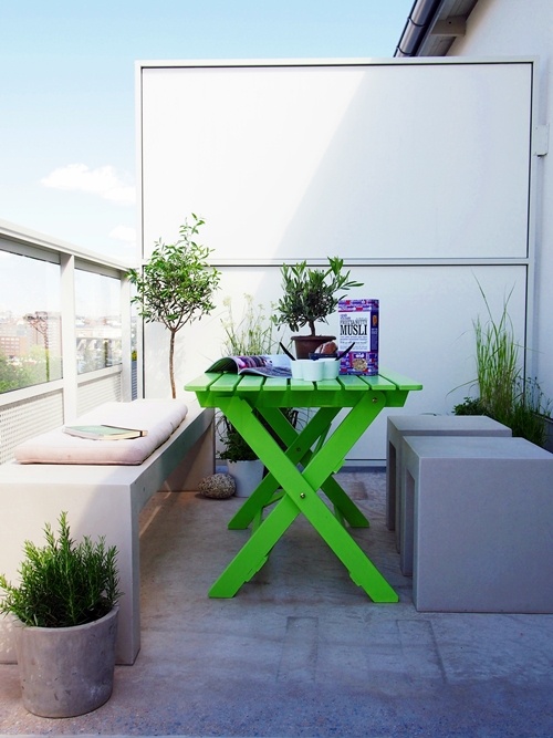 a minimalist Scandi balcony with concrete stools and a bench, a neon green wooden table and potted greenery and blooms