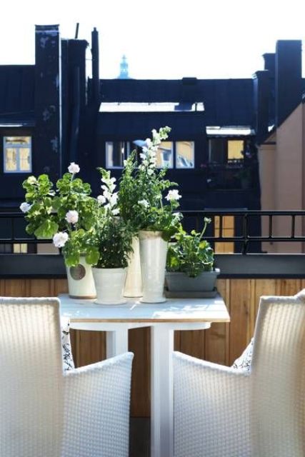 a tiny Nordic balcony with a white table and white wicker chairs, with potted blooms and greenery and a lovely view