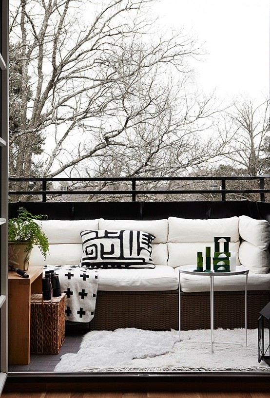 a Nordic balcony with a wicker sofa with white upholstery, a wood and wicker side table, greenery and a glass side table
