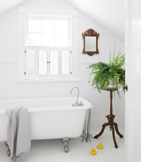 a clean white bathroom with shutters on the window, a vintage tub, a wood frame mirror and a stained plant stand with greenery to refresh the space
