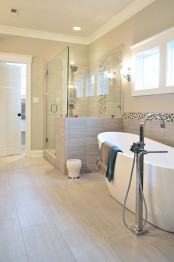 a stylish modern bathroom clad with sandy tiles, with a shower space with half walls and a lovely bathtub is filled with light