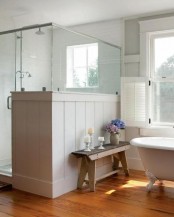 a neutral farmhouse bathroom with planked walls and a shower space, with a pony wall and a vintage bathtub plus a bench