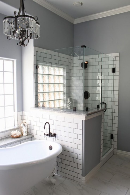 a grey bathroom with white subway tiles, shower space with a half wall, a crystal chandelier and a vintage bathtub