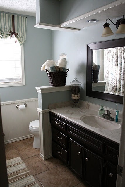 a neutral farmhouse bathroom with light blue walls, a half wall that separates the toilet zone and a dark wooden vanity