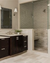 a neutral farmhouse bathroom with a half wall and grey tiles in the shower, a dark stained vanity and a large mirror