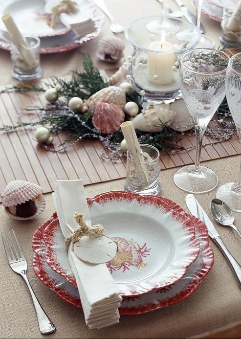 a beach Christmas tablescape with a table runner, coral beach-inspired printed plates, seashells, pearls, pillar candles and napkin rings with seashells