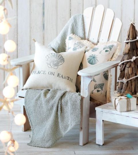 a neutral and pastel beach Christmas nook with a white planked chair, a light blue blanket, printed pillows and a garland