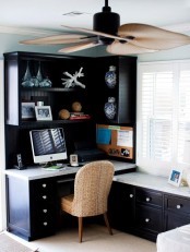 a dark beach home office with a single shelving unit with open and closed storage compartments, a desk and a rattan chair, a large fan on the ceiling