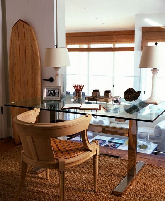 a lovely beach home office done in neutrals, with a glass desk, a rattan chair, a jute rug and a surf board, with shades on the windows