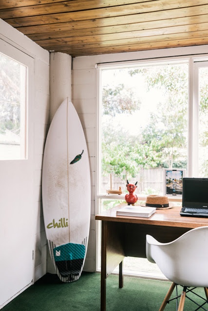 a modern beach home office with a wooden ceiling, a lovely view of outside, a desk and a chair plus a surf board