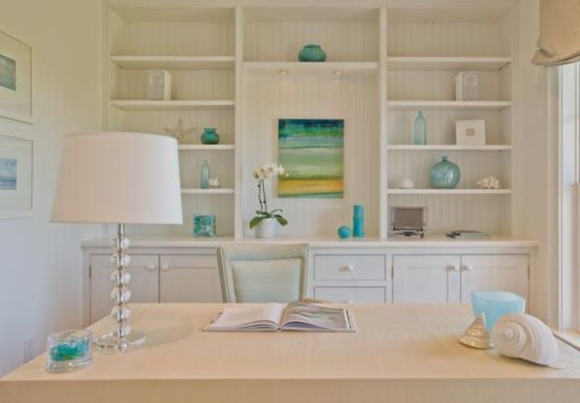 a neutral coastal home office with built in shelves, a desk, a lamp and touches of aqua and blue