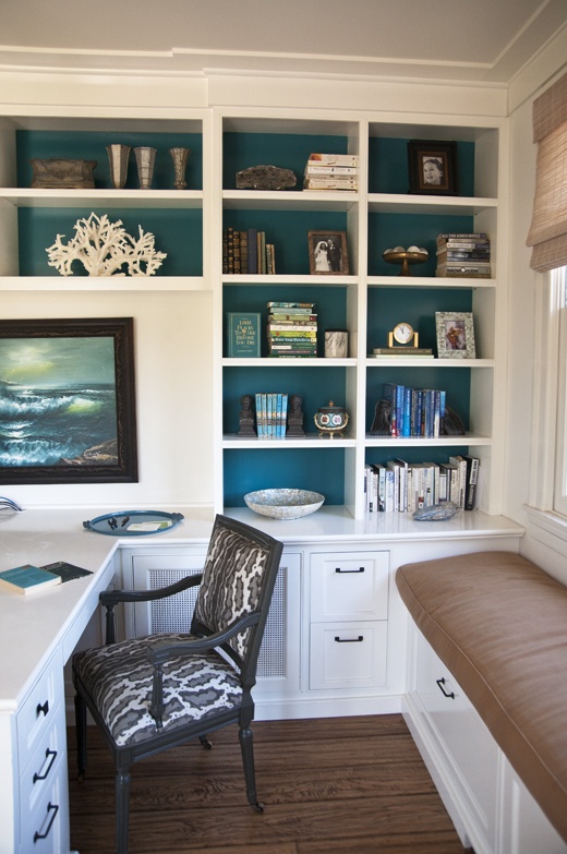 a bold beach home office with teal walls, a white storage unit with open shelves and a built-in desk plus a windowsill upholstered bench is a cool space to be