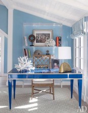 a cool beach or coastal home office with light blue walls, a bold blue desk, a gilded rattan chair, a sheer shelving unit and pretty maritime accessories