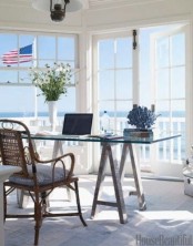 a neutral home office with glazed walls with a beach view, a glass desk and a rattan chair plus greenery and blooms