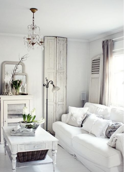 a white shabby chic living room with a vintage door as decor, white seating furniture, a glass storage cabinet, a crystal chandelier, blooms and branches and a floor lamp