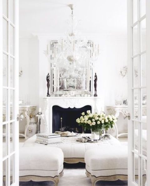 a white vintage living room with a chic non-working fireplace, a large low ottoman and poufs, a crystal chandelier and a large mirror is amazing