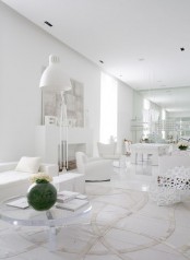 a modern white living room with a faux fireplace, soft white seating furniture, a clear glass table with a green vase, a floor lamp and pretty artwork