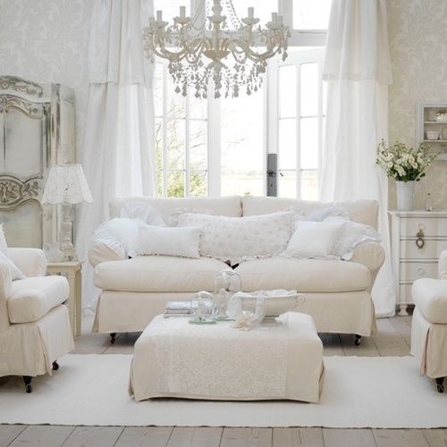 a white vintage living room with neutral seating furniture, a low ottoman, a chic chandelier, a white sideboard, a whitewashed storage unit feels very elegant