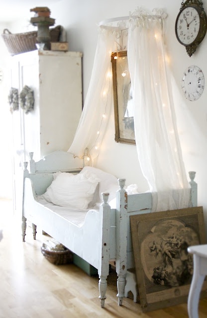 a neutral shabby chic kid's room with a pastel blue bed, white furniture, a canopy with lights and some vintage decor