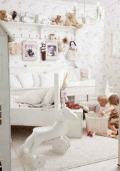 a white shabby chic kid’s room with white furniture and a chandelier, floral wallpaper, a shelf with toys and baskets for storage