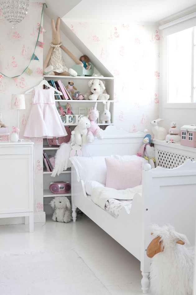 a white and blush shabby chic kid's room with floral wallpaper, chic white furniture, built in shelves and lots of toys and a crystal chandelier over the space