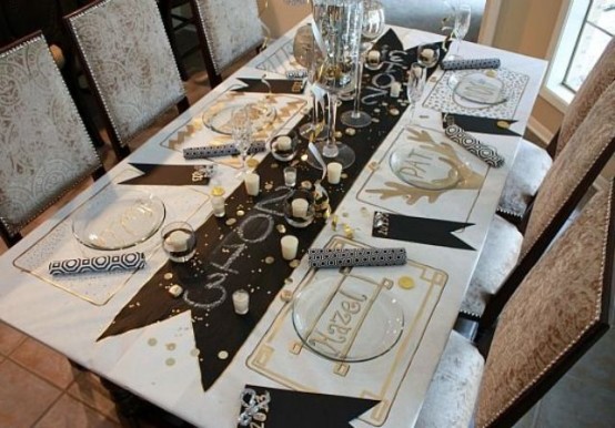 a black, gold and white NYE party tablescape with a table table runner and napkins, printed placemats and clear chargers, candles and gold rhinestones