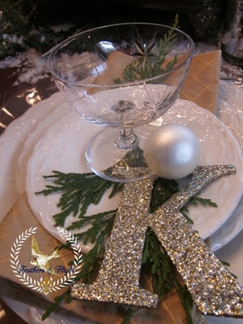 a NYE party place setting with white porcelain, a glass and a pearly ornament, a silver glitter monogram and greenery
