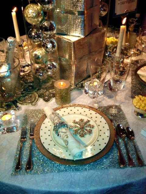 a white and gold NYE party tablescape with a stack of gifts in gold paper, glasses with gold ornaments, a silver placemat and silver cutlery, candles and a gold glitter snowflake