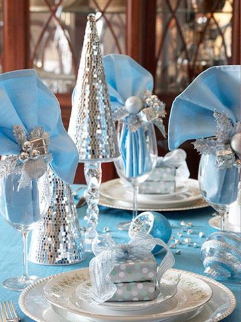 a pastel blue and silver NYE party tablescape with printed white porcelain, a gift with a bow on top, glasses with pastel blue napkins and silver ornaments