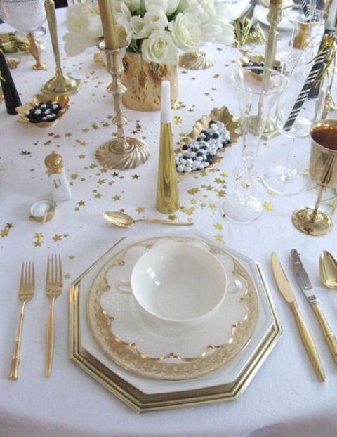 a gold and white NYE tablescape with white blooms, gold candleholders and tall and thin candles, porcelain with gold touches, gold stars on the table