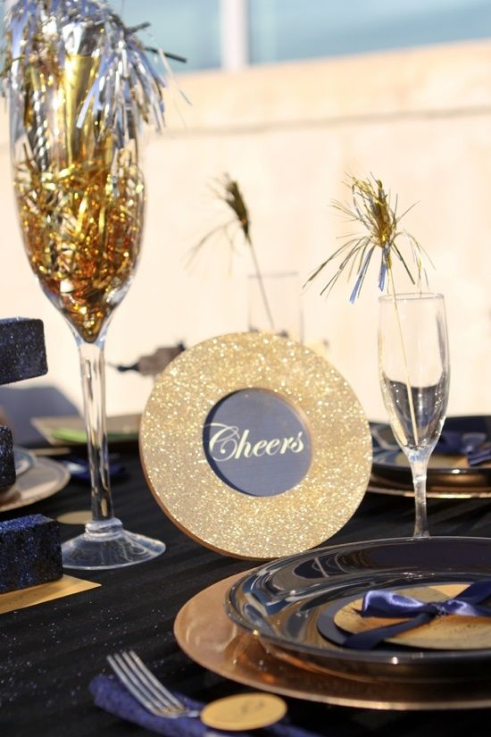 a black, midnight blue and navy NYE tablescape with gold chargers and black plates, a gold glitter sign, some gold cutlery and gold tinsel in glasses